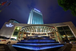 A hotel with a modern glass facade, a prominent entrance with a glass canopy, and a blue-lit water feature in front. - Minshan Hotel - Chengdu (Chengdu)