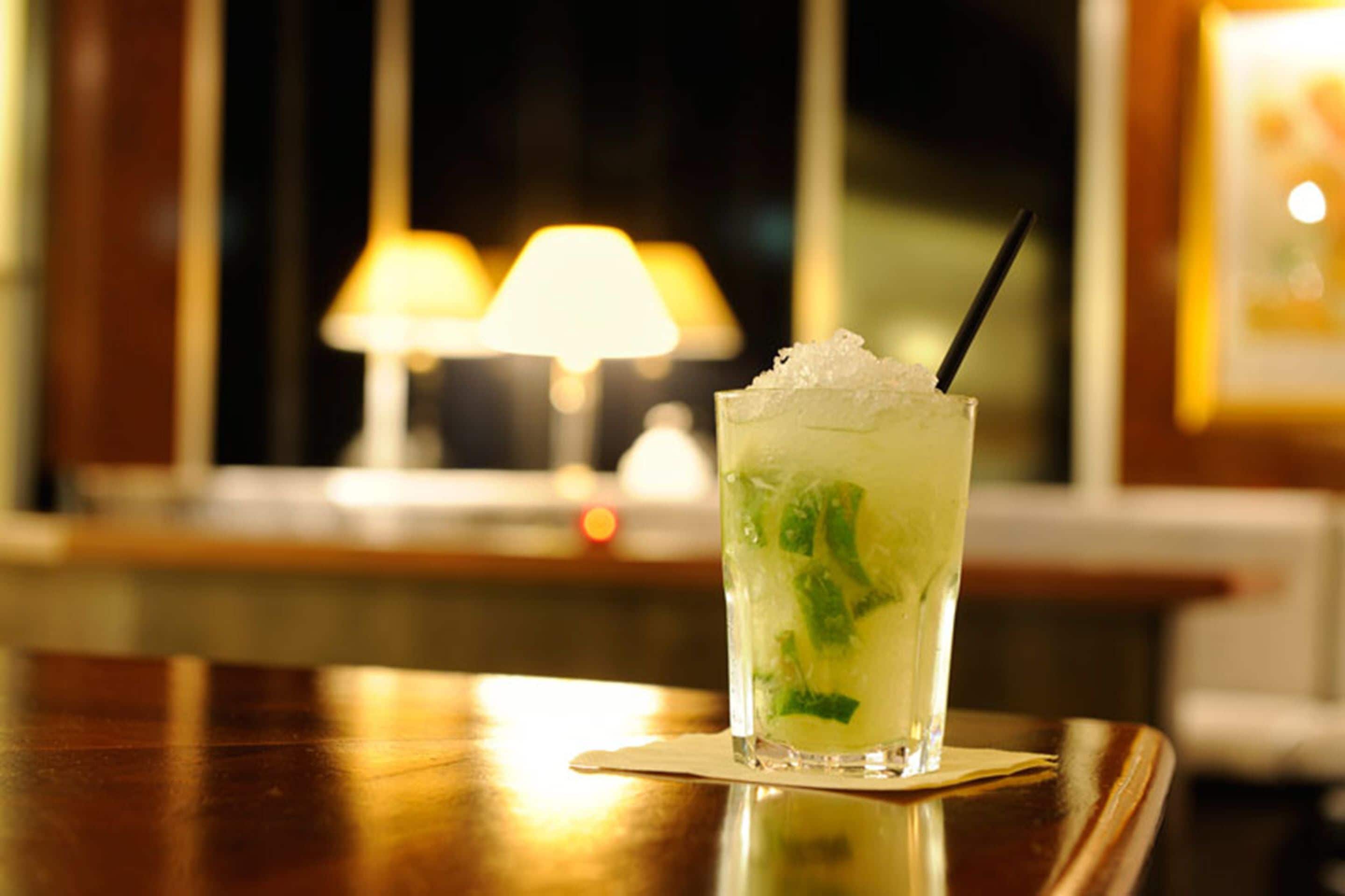 A cocktail glass with ice and mint leaves on a coaster, placed on a wooden table with a lamp in the background.