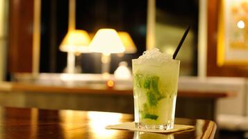 Un vaso de cóctel con hielo y hojas de menta sobre un posavasos, colocado en una mesa de madera con una lámpara al fondo.