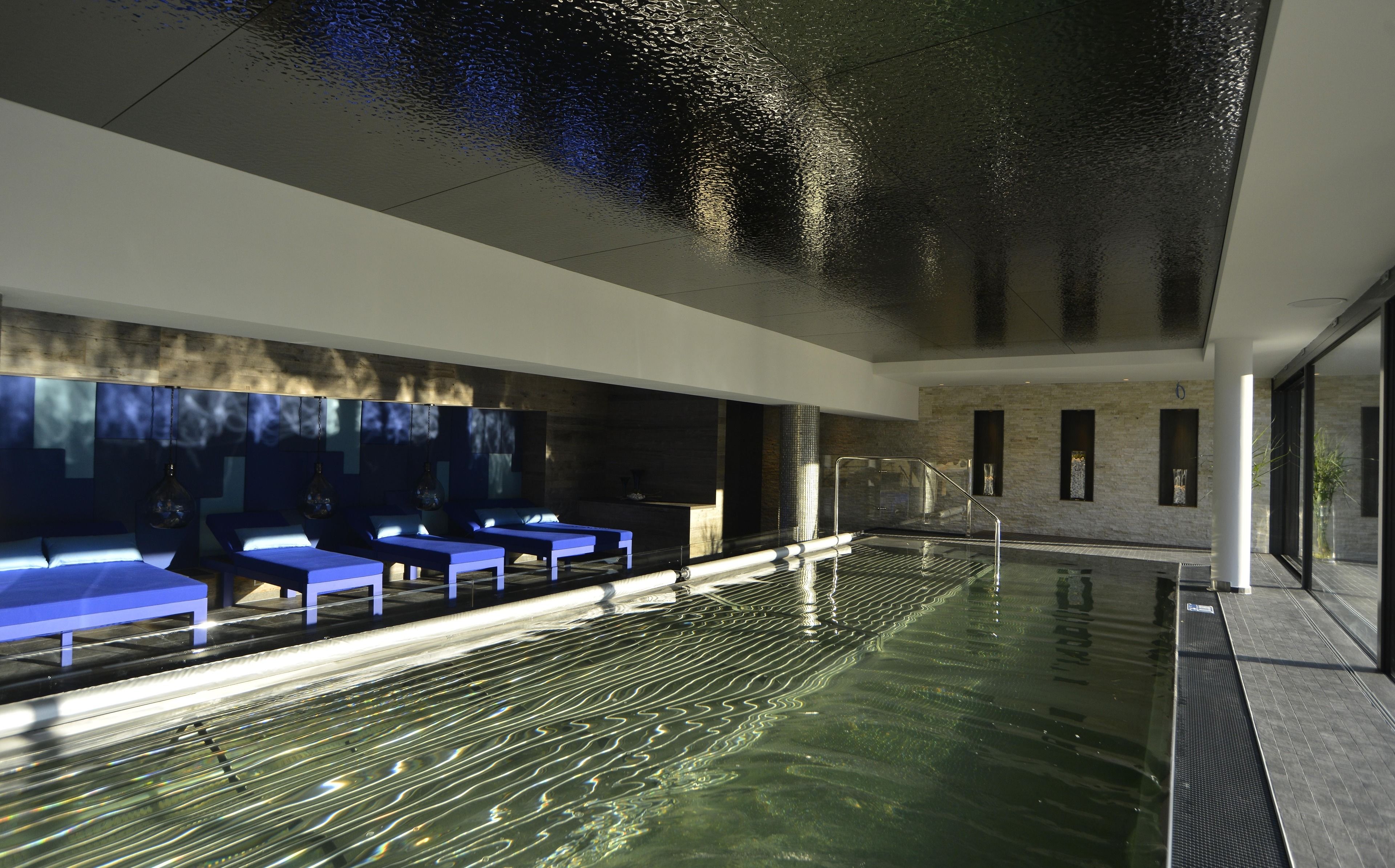 Una piscina interior moderna con agua cristalina, un área de descanso con bancos azules y una pared de piedra con ventanas.