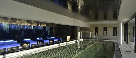 Una piscina interior moderna con agua cristalina, un área de descanso con bancos azules y una pared de piedra con ventanas.