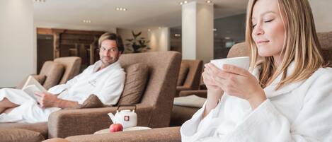 Two people in white bathrobes sitting on reclining chairs in a spa-like setting with a small table between them.