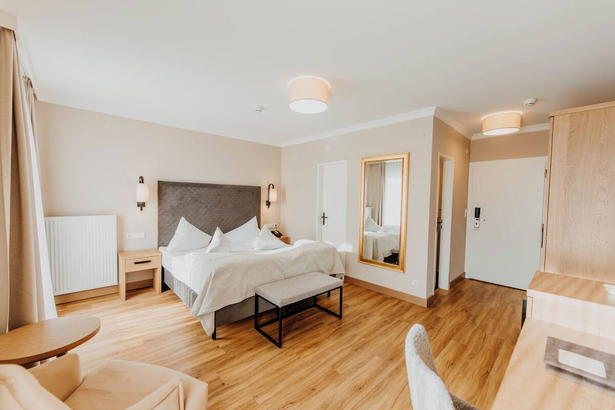 Una habitación de hotel moderna con suelo de madera, una cama grande con ropa blanca, una mesita de noche con lámpara, un espejo y un armario.