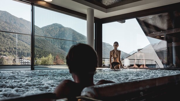 Una persona sentada en un jacuzzi con vistas a montañas a través de grandes ventanales.