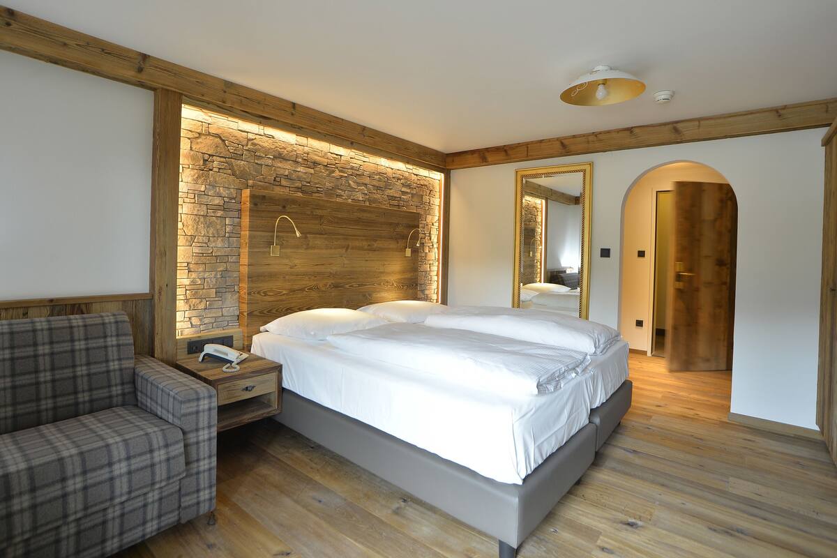 Un dormitorio moderno con una pared de acento de piedra, un cabecero de madera, una cama grande, una mesita de noche con un teléfono y un sofá a cuadros.