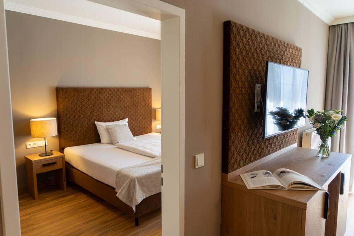 Habitación de hotel con cama, mesita de noche con lámpara, espejo y escritorio con un florero.