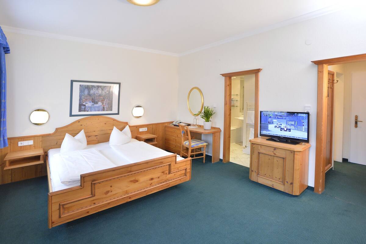 Habitación de hotel con una cama de madera, un escritorio con silla, televisión y baño visible a través de una puerta abierta.