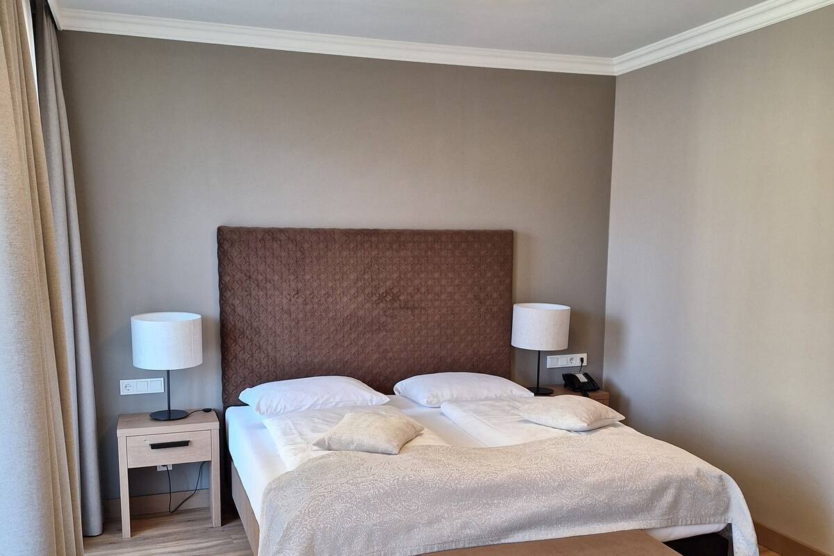 Habitación de hotel ordenada con una cama grande, lámparas de noche, un banco y una lámpara de techo.