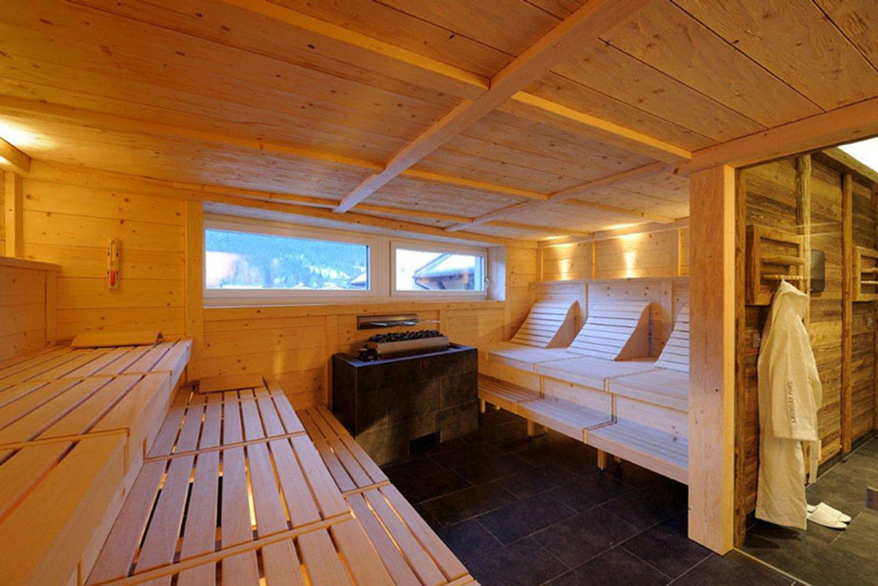 A wooden sauna room with benches and a stone bench, large windows, and a towel rack.
