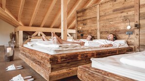 Interior de una cabaña de madera con una cama grande y una más pequeña, ambas con ropa de cama blanca.