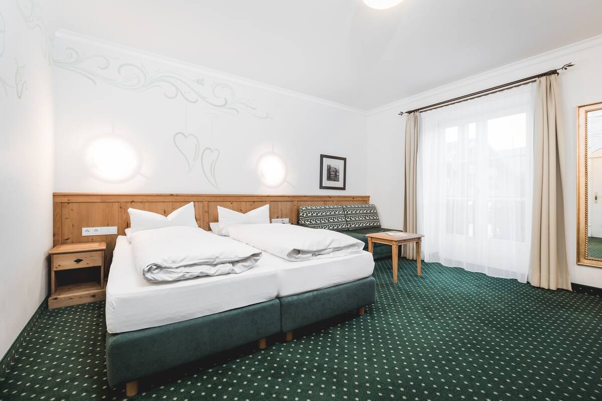 Habitación de hotel con cama doble, mesita de noche de madera, una pequeña mesa, un espejo y un ventanal grande con cortinas.