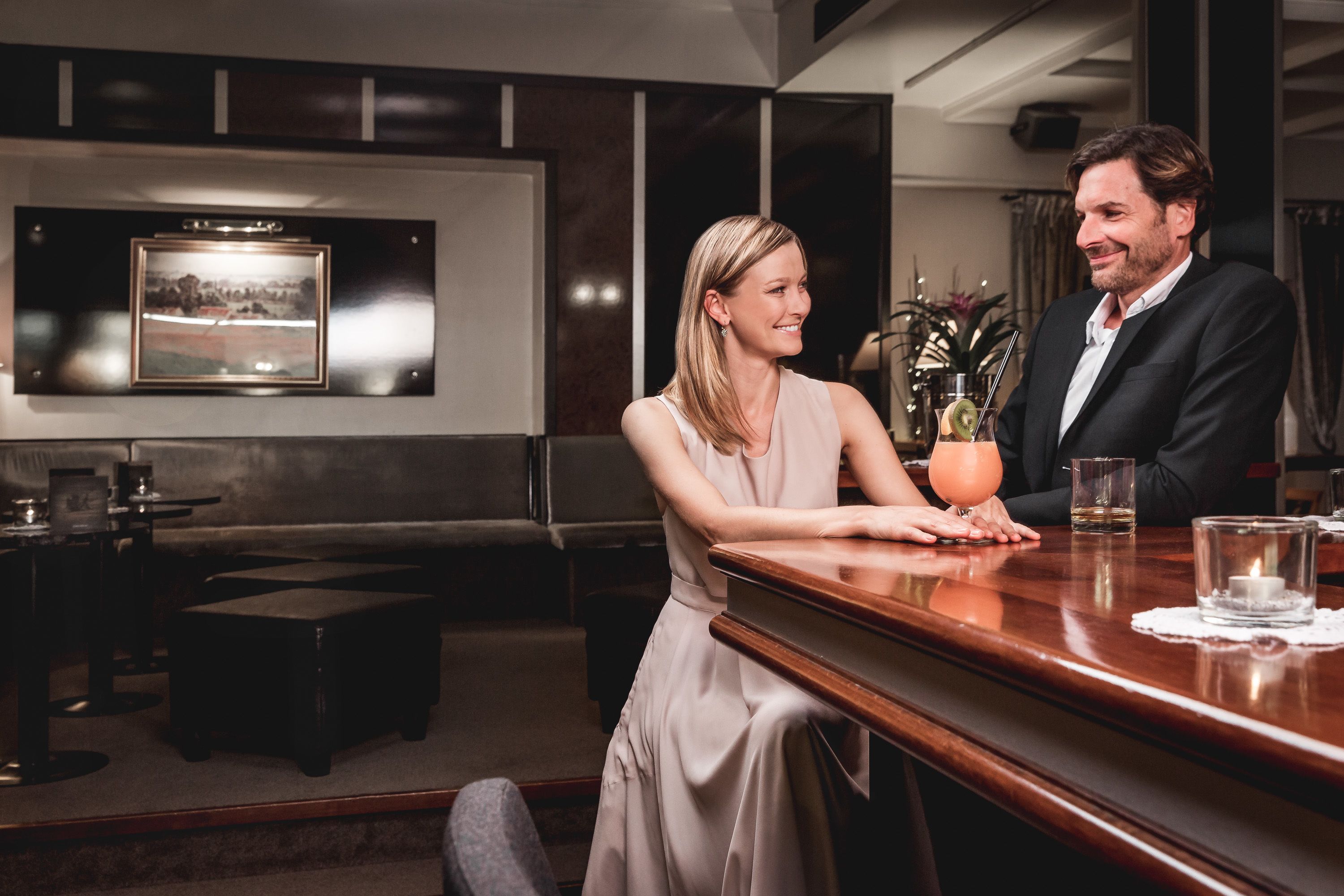A couple seated at a bar in a modern hotel lobby with a framed picture on the wall, a small plant, and a candle on the table.