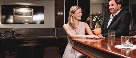 A couple seated at a bar in a modern hotel lobby with a framed picture on the wall, a small plant, and a candle on the table.