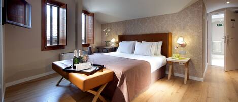 Quarto de hotel com uma cama grande, um banco de madeira, uma pequena mesa e duas luminárias de cabeceira.