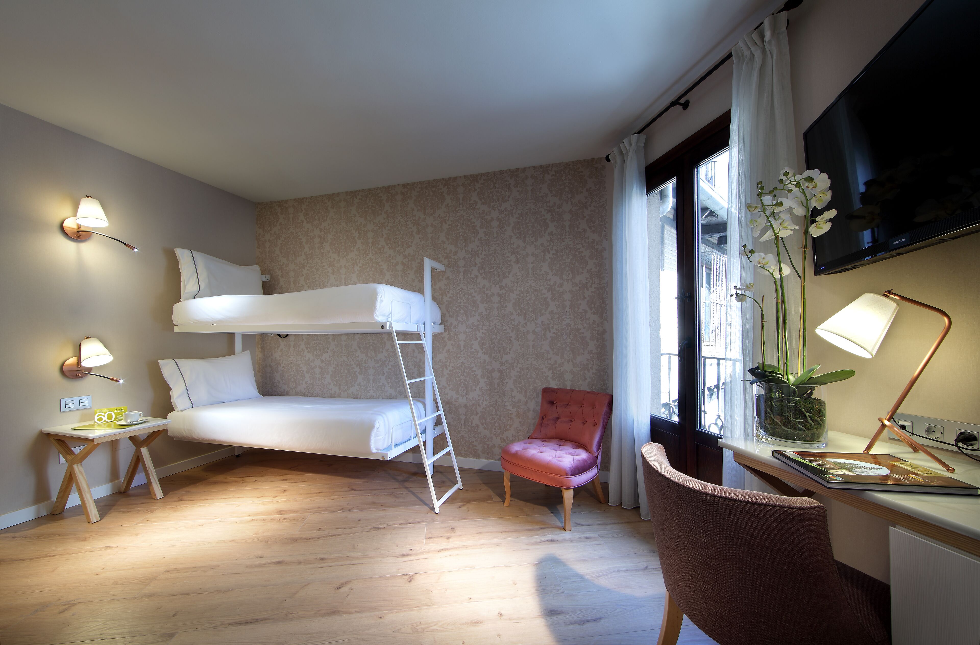Ein kleines Hotelzimmer mit Etagenbett, Schreibtisch mit Lampe, Stuhl, Nachttisch mit Lampe und einem Blumentopf.