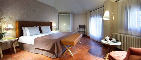 Habitación de hotel con cama, banca, silla, mesita de noche, lámpara y vista al exterior a través de cortinas.
