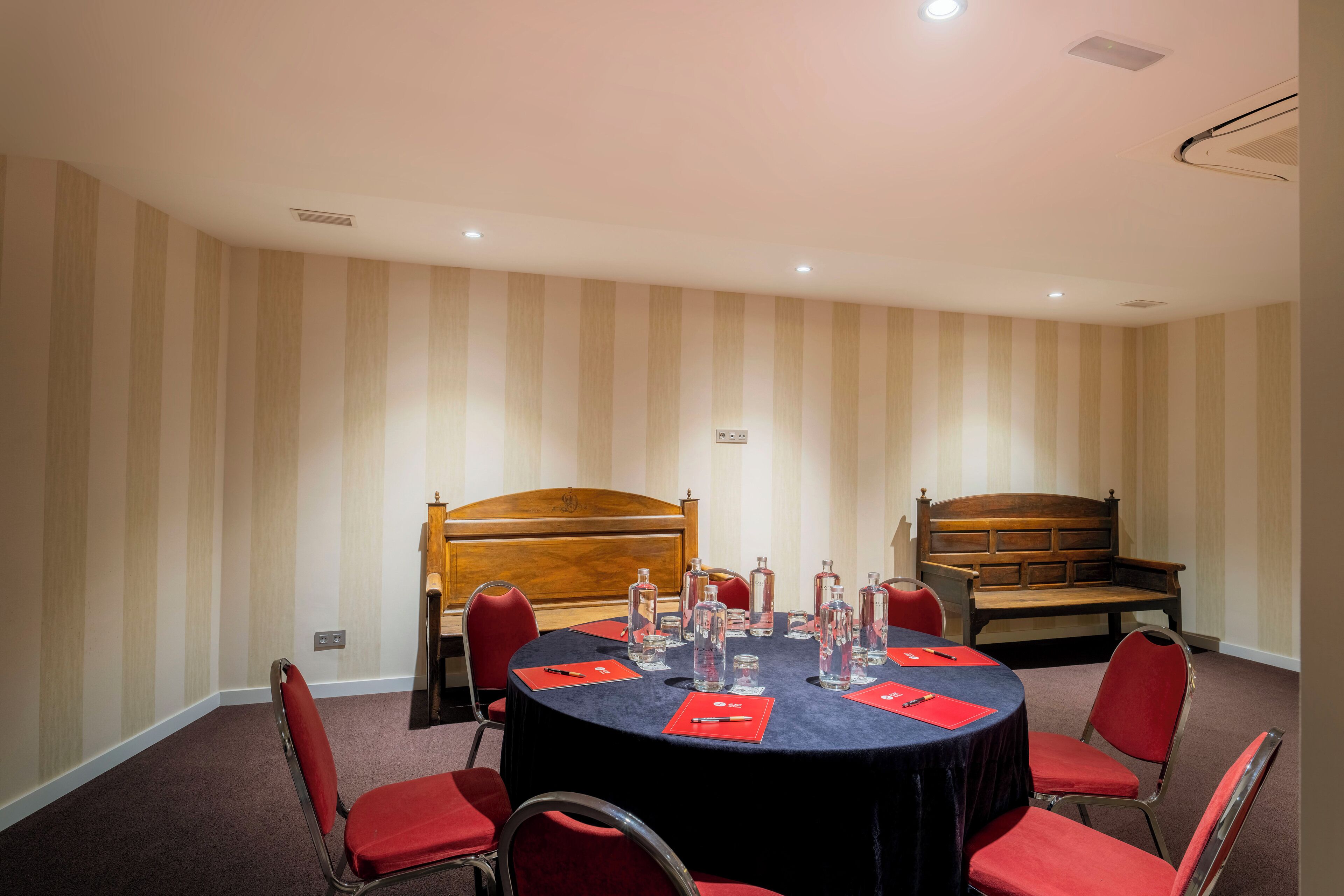 Un salón de conferencias con una mesa redonda, sillas rojas, un banco de madera y papel tapiz a rayas.
