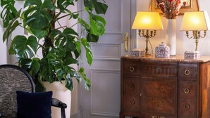 A room with a potted plant, a chair with a cushion, a wooden dresser with drawers, and two wall-mounted lamps. - Le Relais Madeleine (Paris)