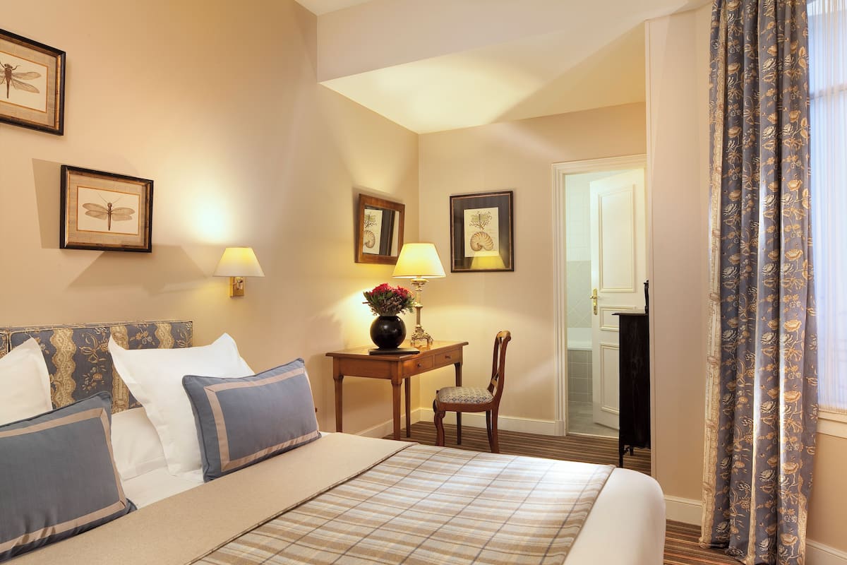A hotel room with a bed, a desk with a lamp, and framed artwork on the walls.