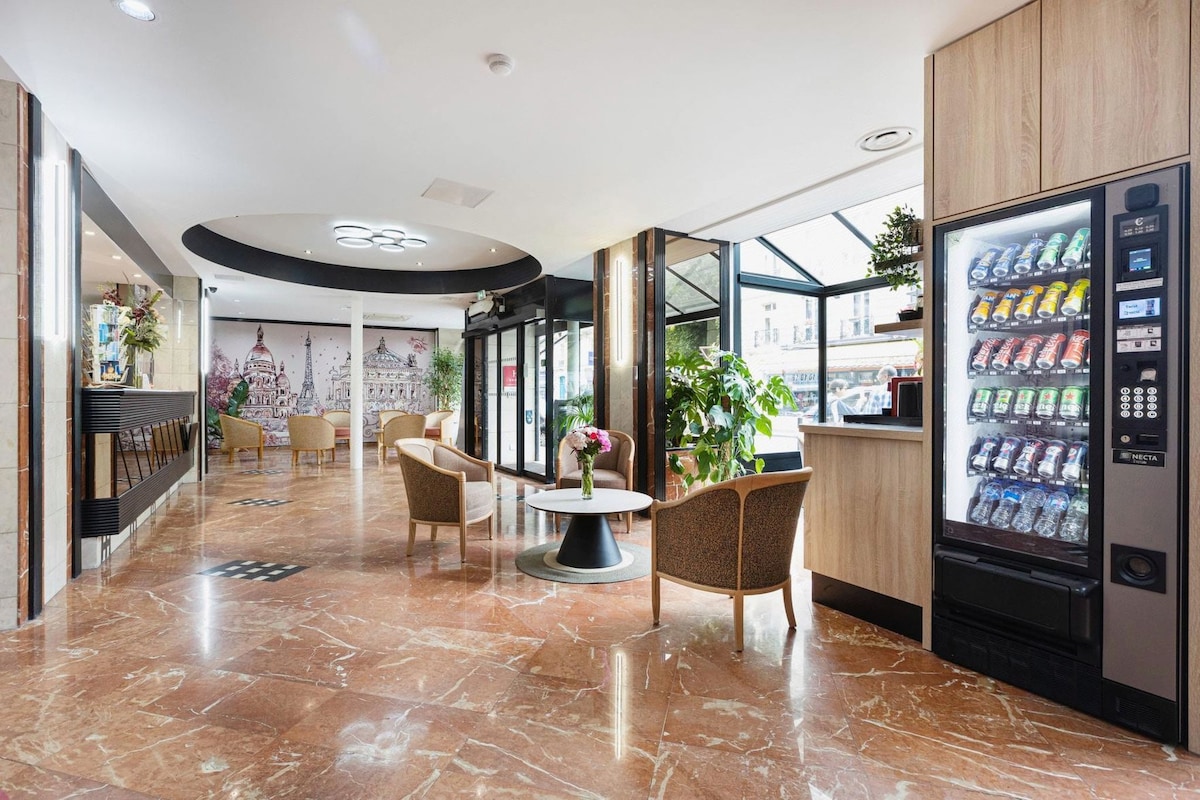 A hotel lobby with marble flooring, seating area with chairs and a round table, a wall mural, and a vending machine.