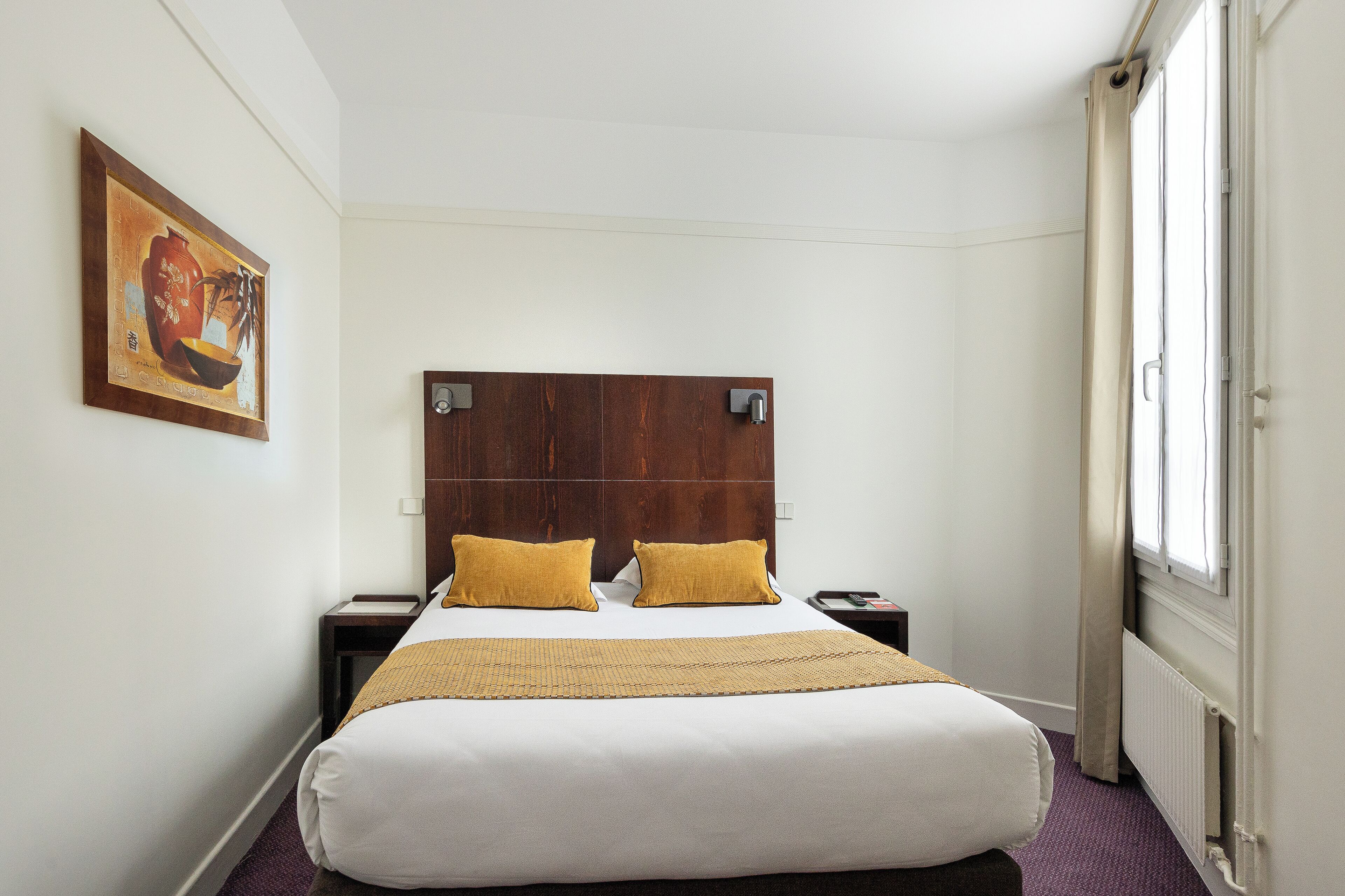 A hotel room with a large bed, two pillows, a headboard, a nightstand, a lamp, a painting on the wall, and a window with curtains.