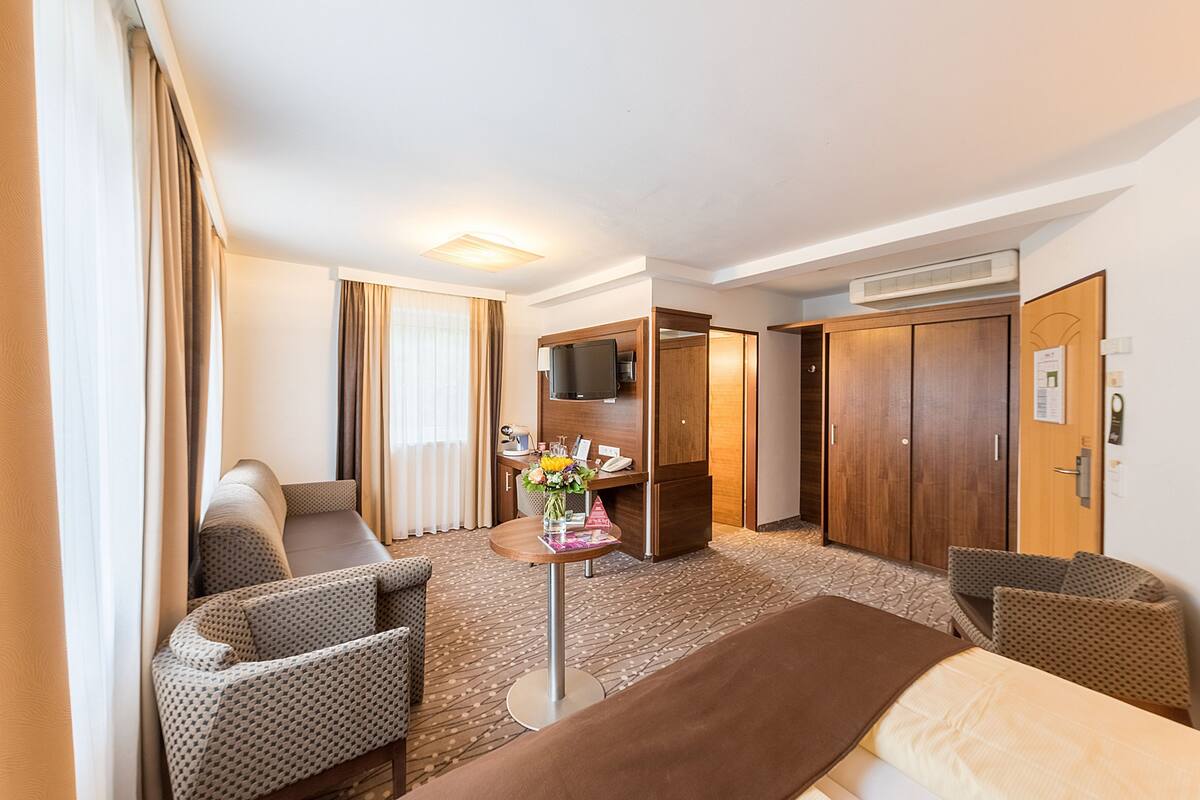 A hotel room with a bed, two armchairs, a small table with a vase of flowers, a television, and a wardrobe.