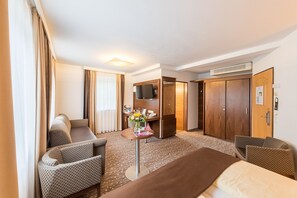 Hotellihuone, jossa on sänky, kaksi nojatuolia, pieni pöytä, jossa on maljakko täynnä kukkia, televisio ja vaatekaappi.