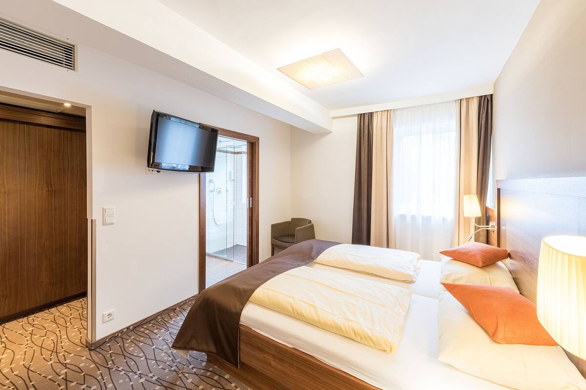 A hotel room with a bed, a television mounted on the wall, a window with curtains, and a lamp on the side table.