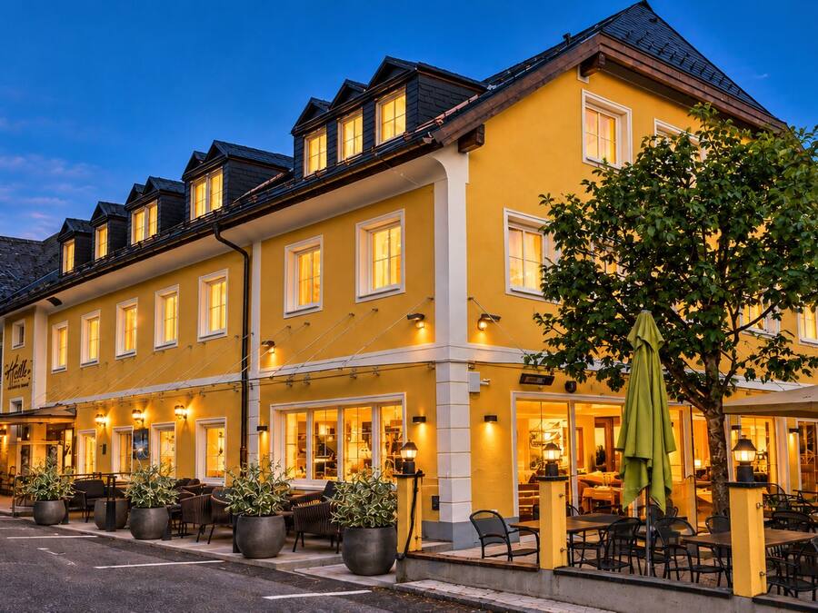 A two-story building with a yellow facade, multiple windows, outdoor seating area with tables and chairs, and a tree in the foreground.
