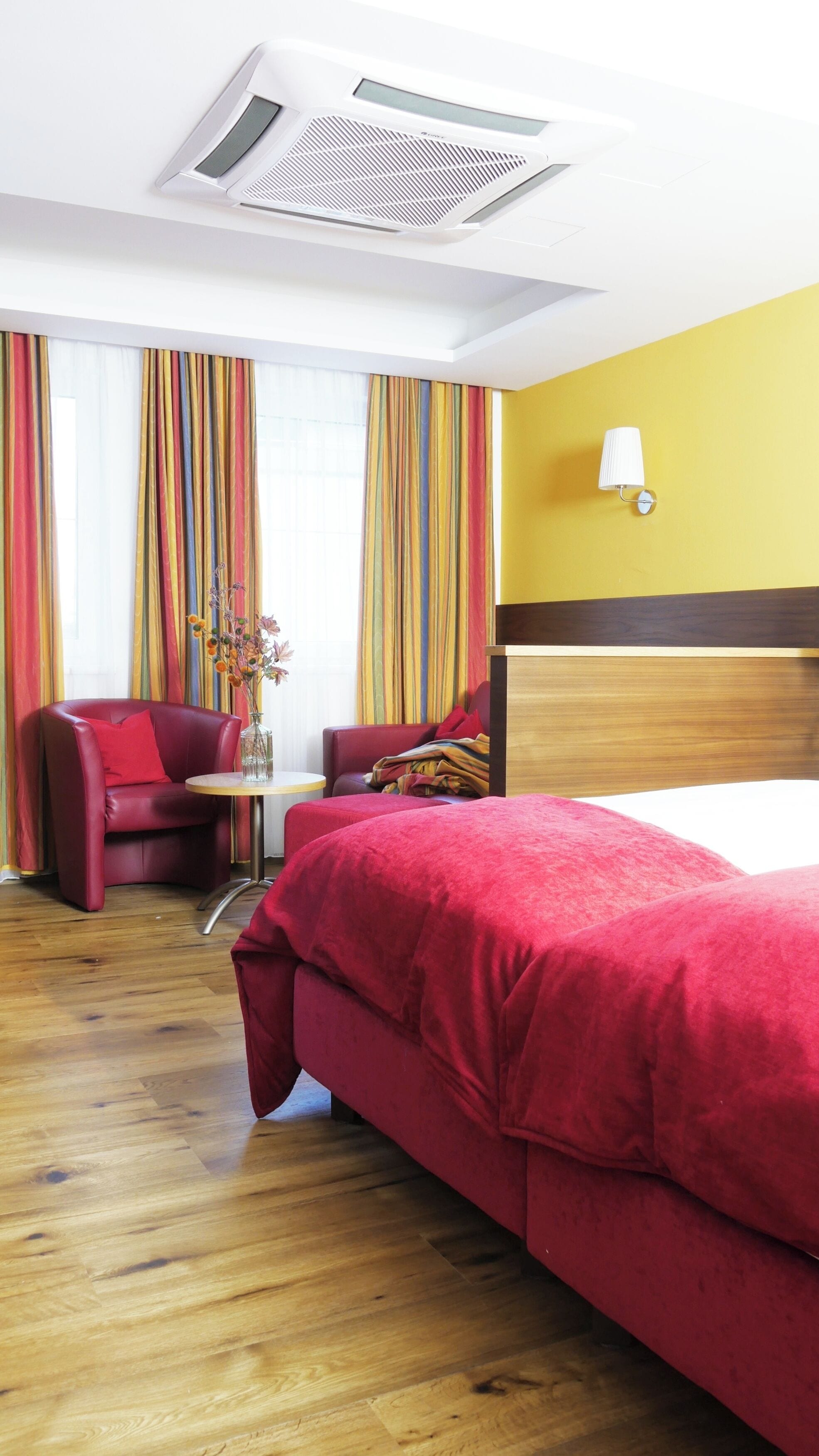 A hotel room with a wooden floor, a bed with red bedding, a red armchair, a small table with a vase of flowers, and a window with colorful curtains.