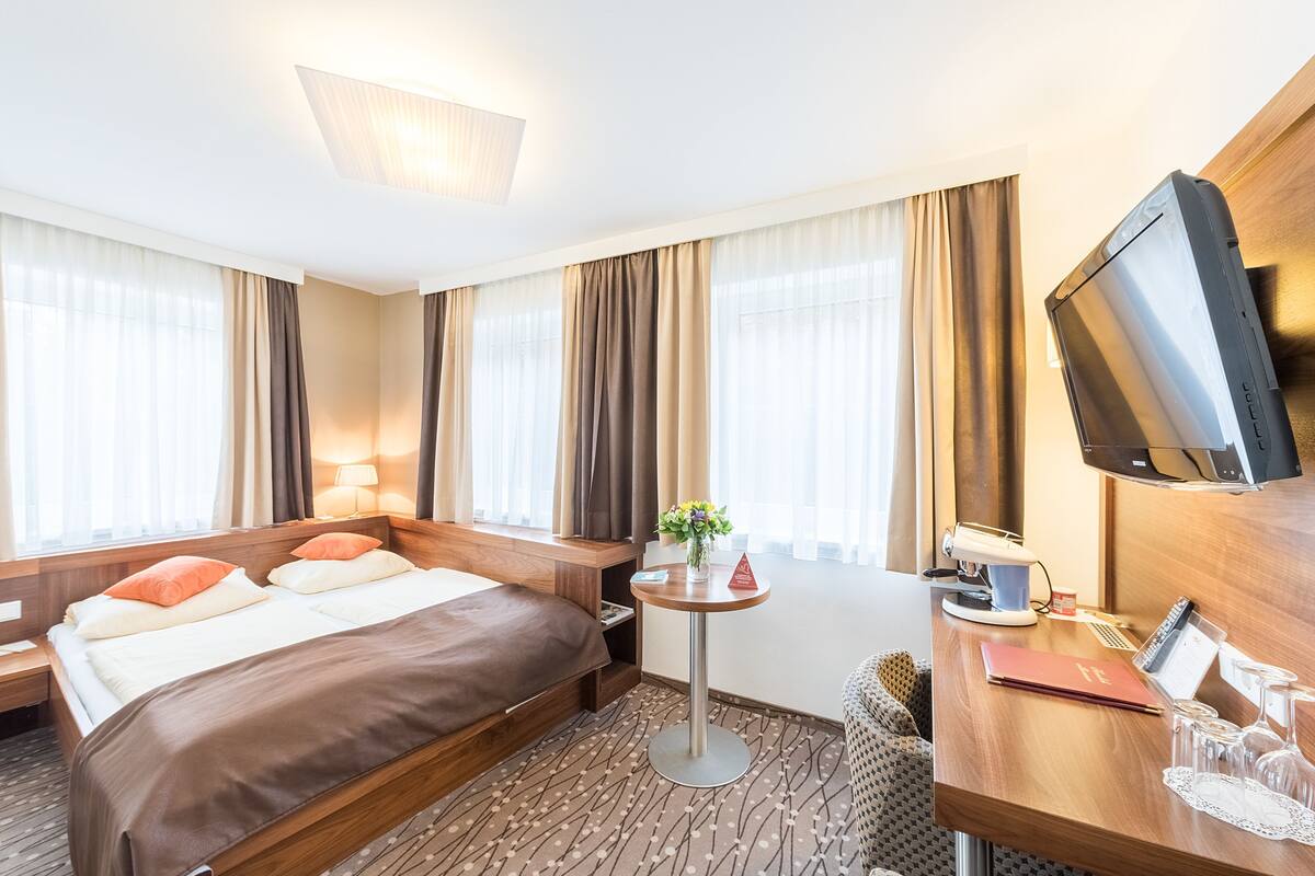 A hotel room with a bed, bedside table, TV, desk, and a small table with a plant.