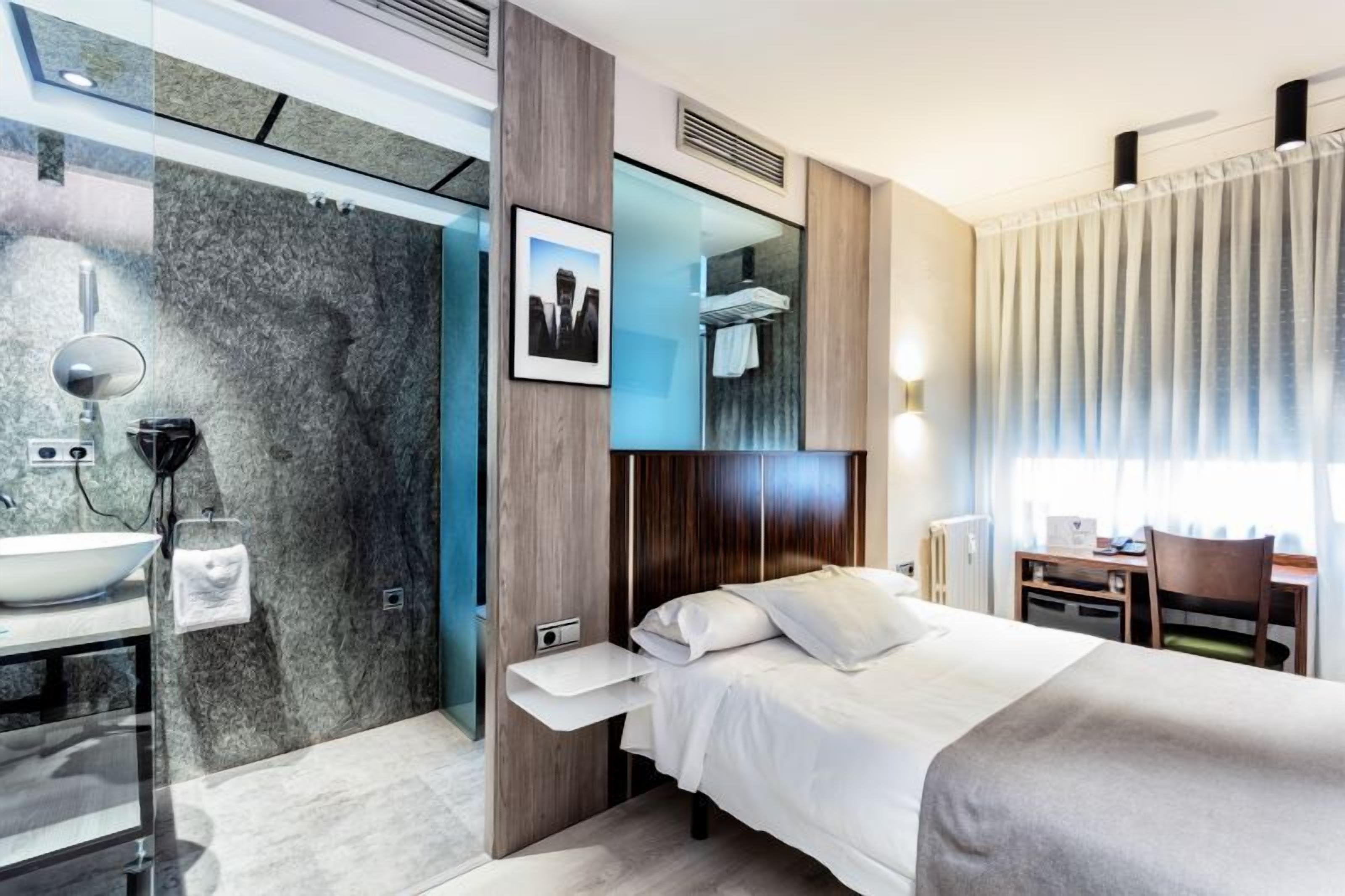 Une chambre d’hôtel moderne équipée d’un lit, d’une table de chevet et d’une salle de bain avec un lavabo et un miroir.