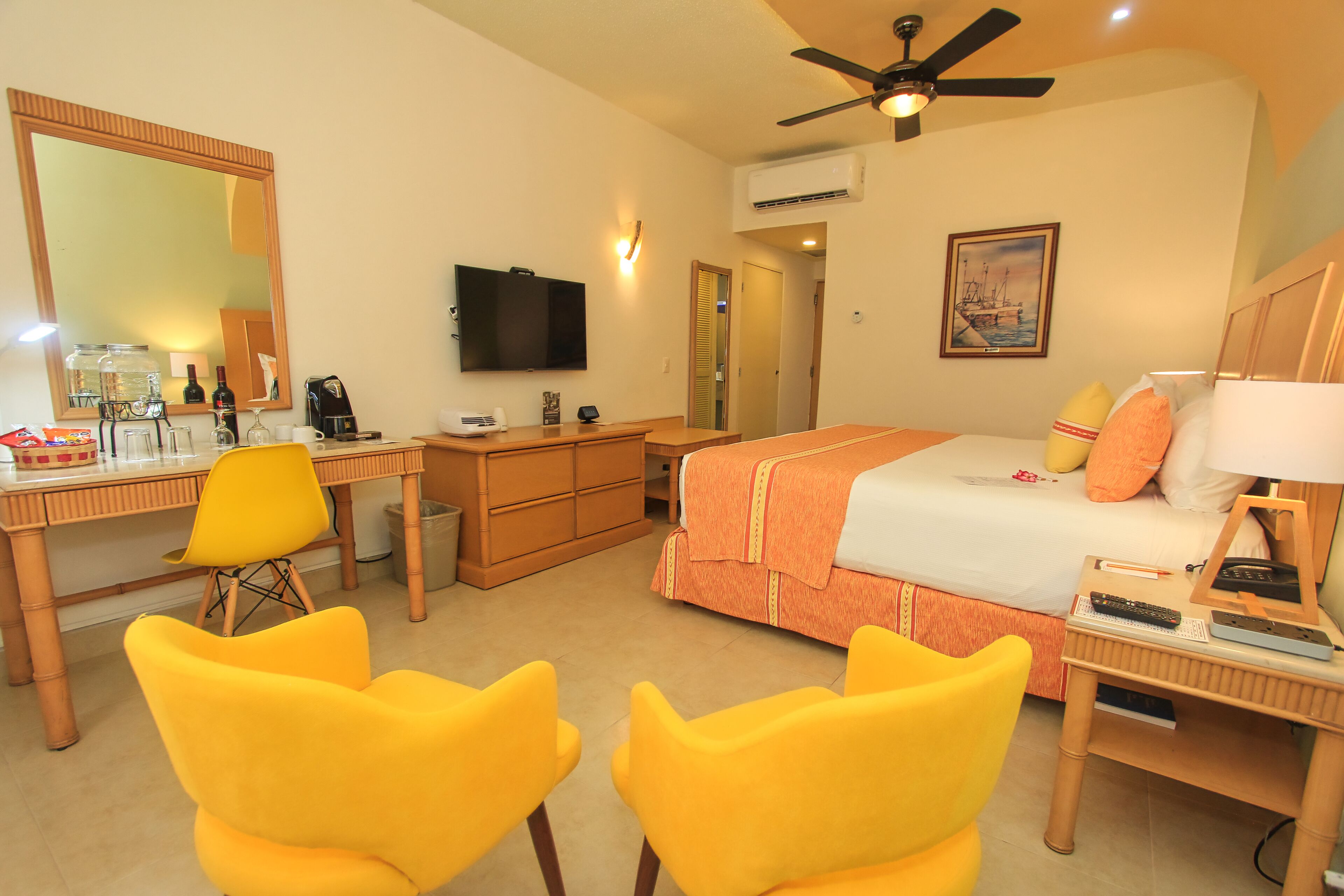 A hotel room with a bed, desk, chairs, a television, and a ceiling fan.