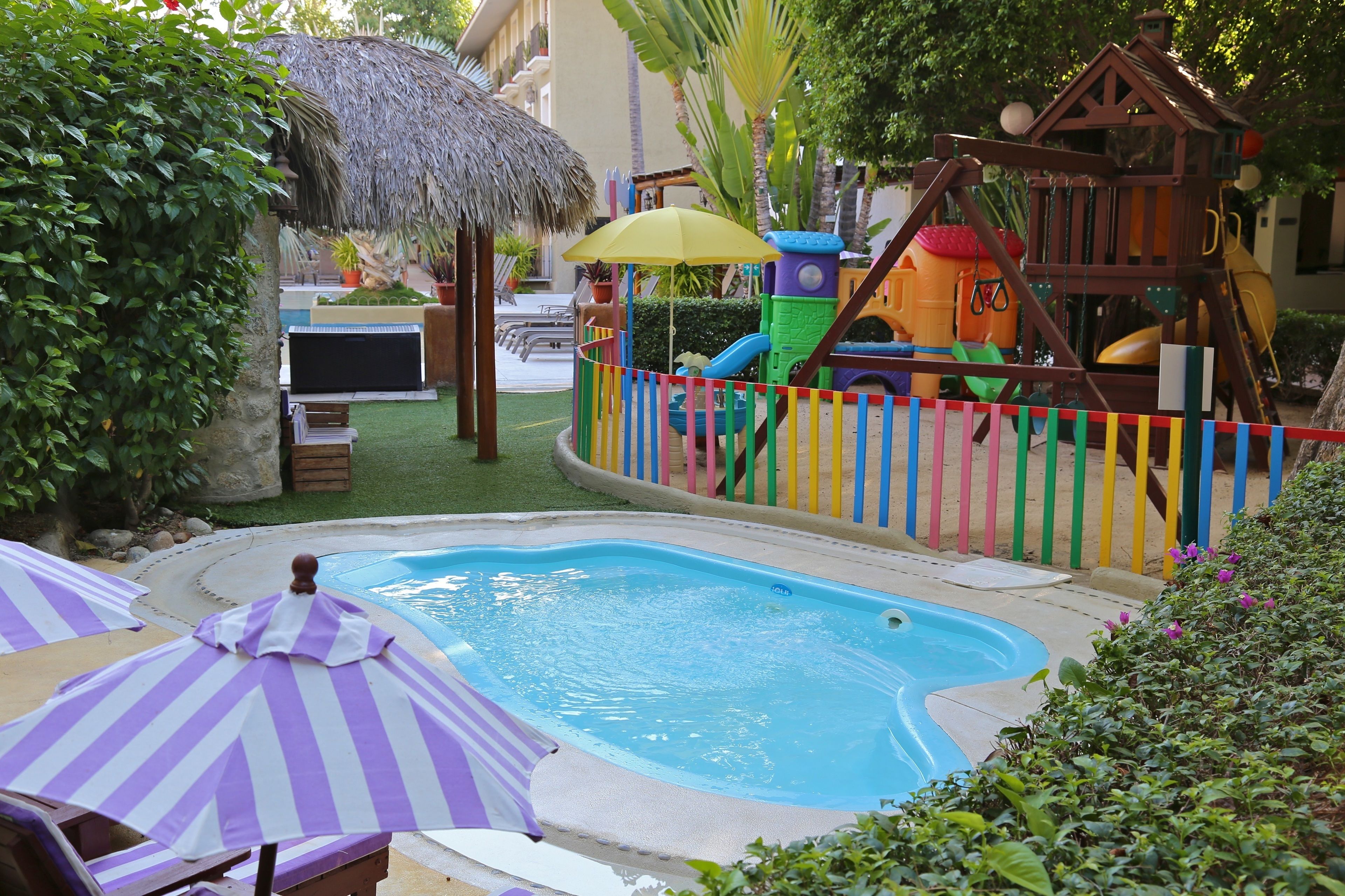 Una piscina con plataforma de madera, rodeada por una cerca colorida y un parque infantil.