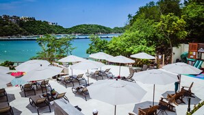 A seaside resort with outdoor seating and umbrellas, overlooking a clear blue sea and a forested hillside.