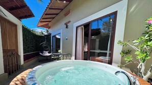 A hot tub in a sunlit outdoor area with a wooden deck, a hanging chair, and a glass door leading to a bathroom.