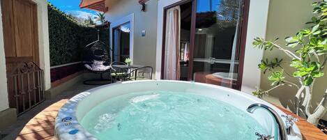 Een hottub in een zonnig buitengebied met een houten terras, een hangstoel en een glazen deur naar een badkamer.