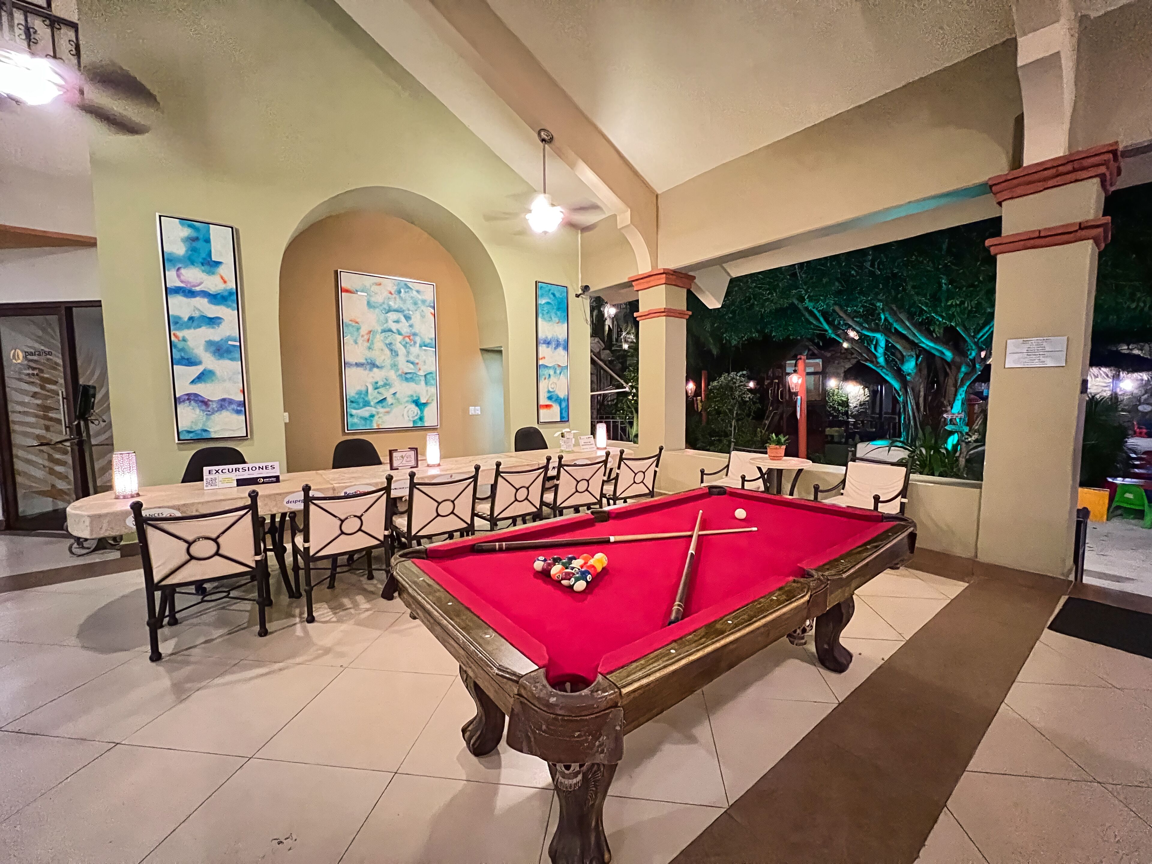 A pool table with a red felt surface, surrounded by chairs and tables in a room with tiled flooring and a high ceiling.