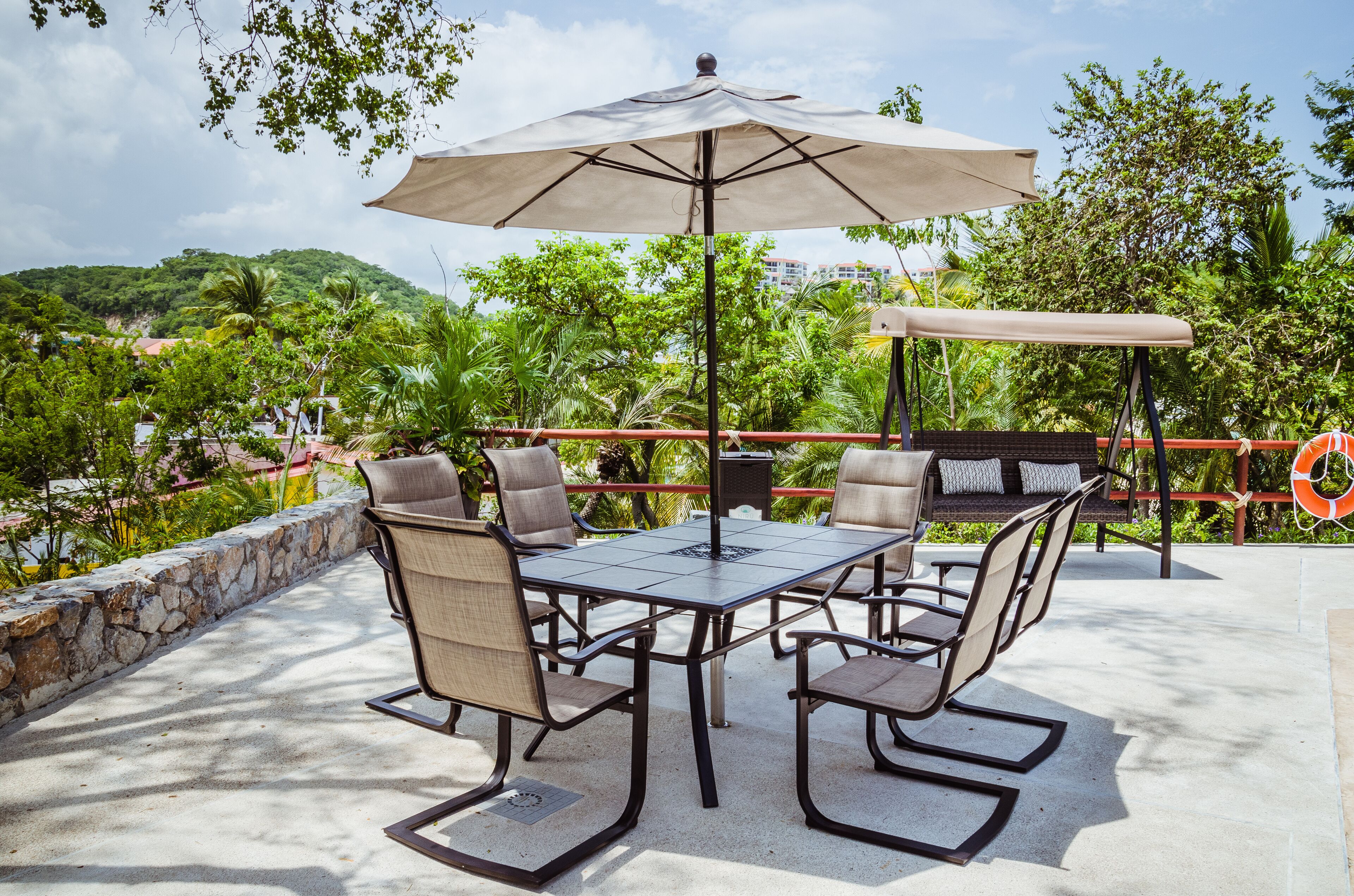 An outdoor patio with a table and chairs, a large umbrella, and a view of greenery and buildings in the background.