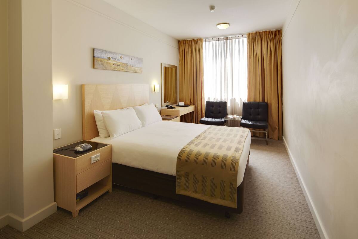 A hotel room with a bed, bedside table, desk, chair, and a large window with curtains.