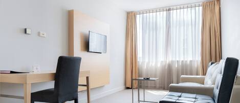 Una habitación de hotel moderna con un escritorio de madera, una silla negra, un sofá color beige y un televisor montado en la pared.