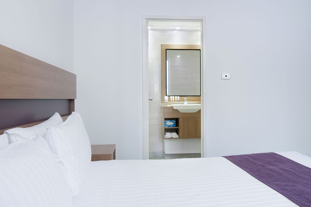 A hotel room with a bed, a nightstand, a bathroom with a mirror and sink, and a wooden headboard.