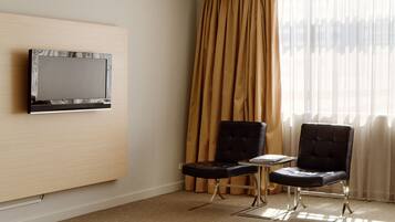 Habitación de hotel con televisor de pantalla plana montado en la pared, una mesita con una silla y un ventanal con cortinas.