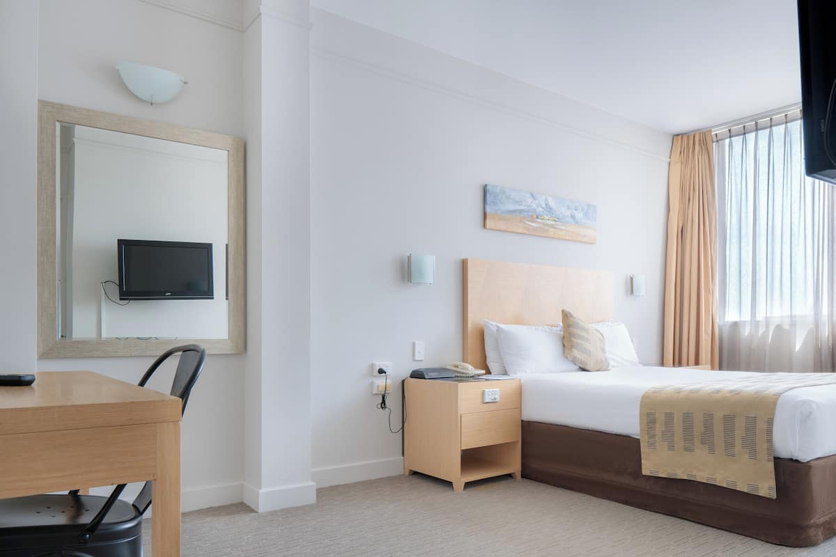 A hotel room with a bed, bedside table, a desk with a chair, a television, and a painting on the wall.