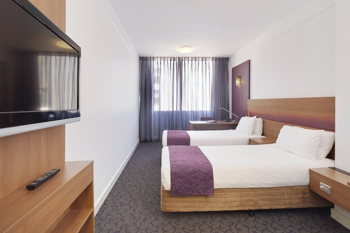 A hotel room with two beds, a TV, a desk, and a window with curtains.