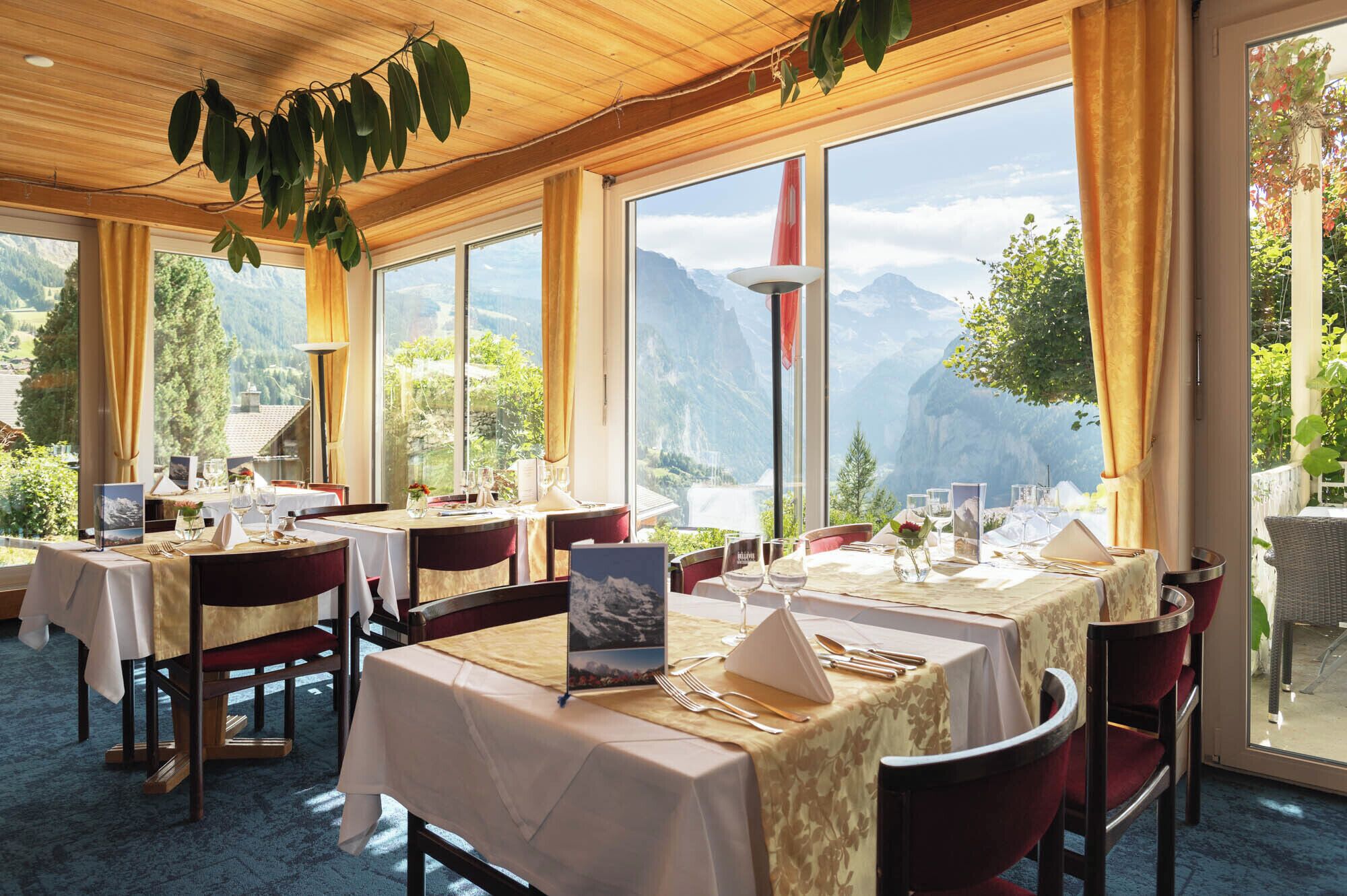 Un restaurante con varias mesas dispuestas para comer, grandes ventanales que ofrecen vistas a la montaña y un techo de madera.