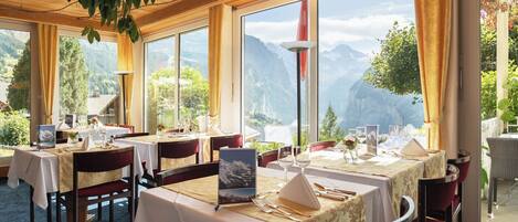 Un restaurante con varias mesas dispuestas para comer, grandes ventanales que ofrecen vistas a la montaña y un techo de madera.