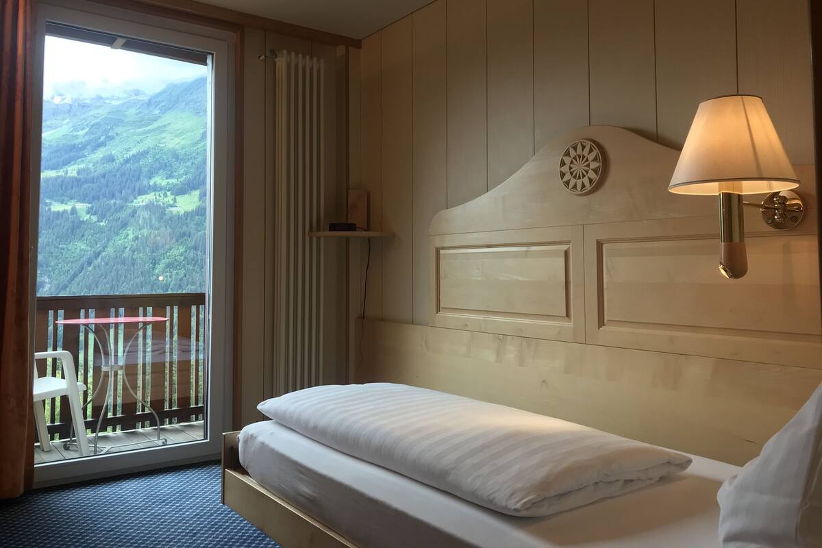 Ein Hotelzimmer mit Bett, Fenster mit Blick auf eine Berglandschaft, einer Wandlampe und einem kleinen Regal.