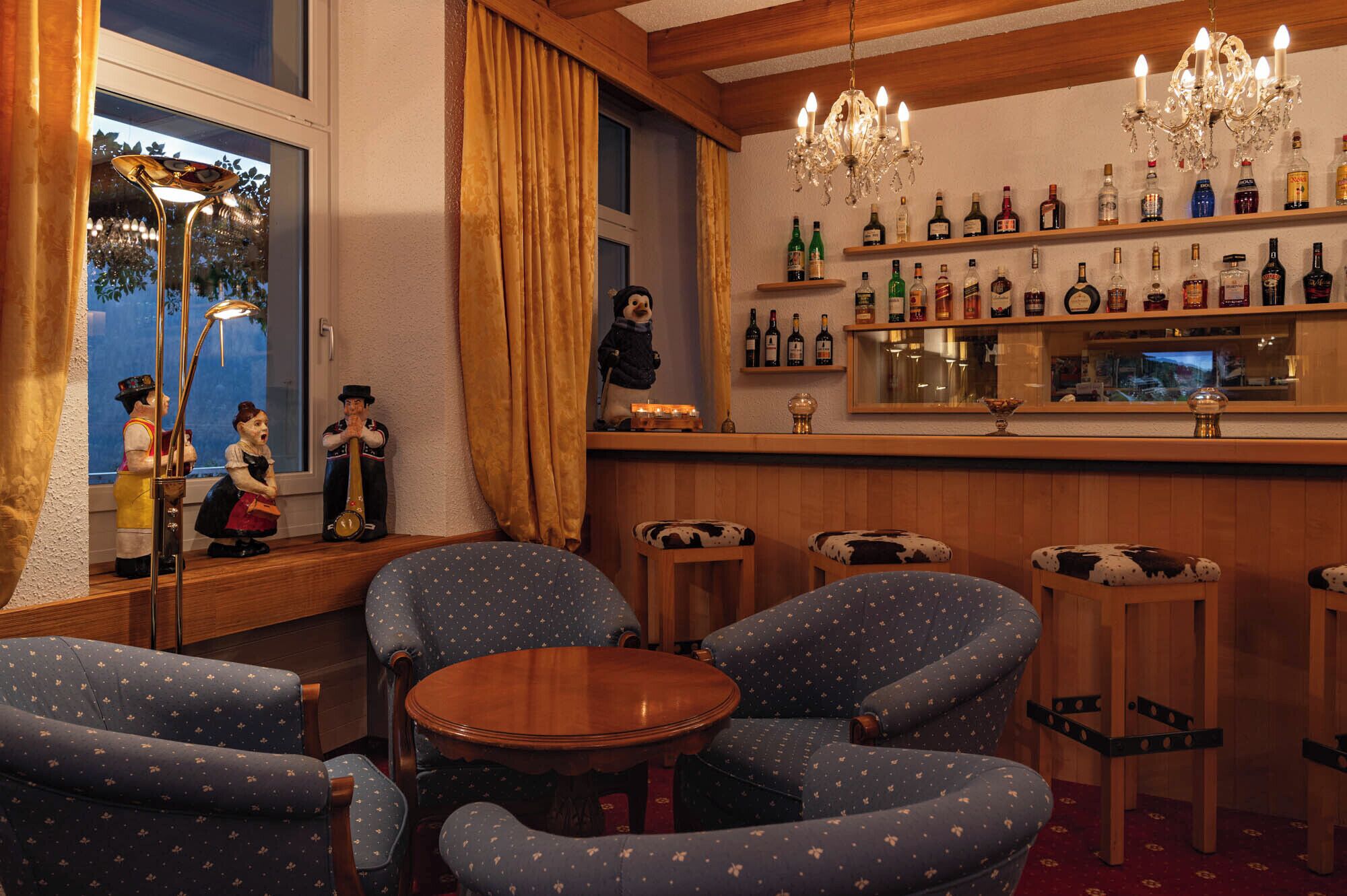 Un espace bar doté d’un comptoir en bois, de plusieurs chaises recouvertes de cuir de vache, d’un lustre et d’une variété de bouteilles d’alcool.