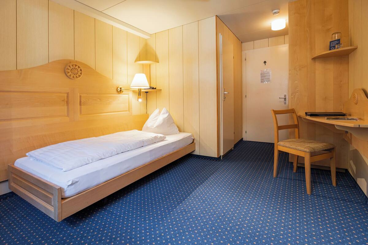 Ein Einzelbett mit Holz-Kopfteil, ein Schreibtisch mit Stuhl, eine Lampe und ein kleines Regal mit Gegenständen.